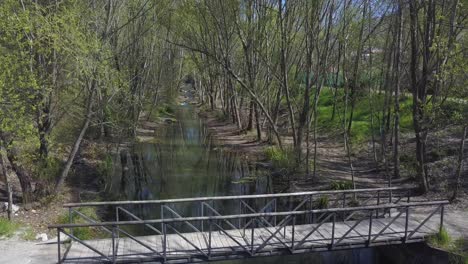 Small-Wooden-Bridge-Over-Shallow-Stream-Between-Woods-At-Daytime