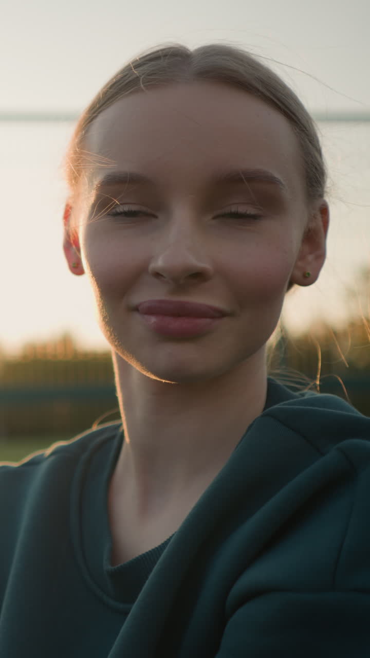 Close-up head view of young white female engaged in outdoor sport activity with focused expression, captured in soft sunlight with blurred background featuring greenery and fencing