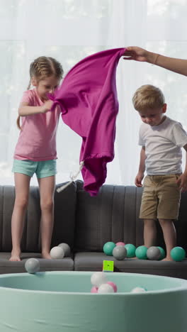 loving mother with daughter and son pours colorful balls from bag into dry pool. toddler boy plays with rubik cube. happy family has fun together at home