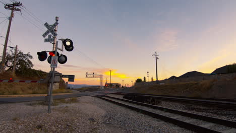 a railway crossing gate's lights flash as it opens after a train has passed and allows cars to go through at sunset.