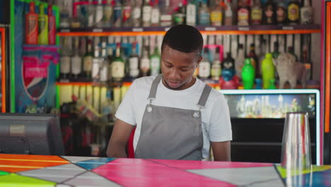 bartender puts ice cubes into glass with scoop in bar. african american man prepares exotic drink for resting guests. young black barman enjoys work routine