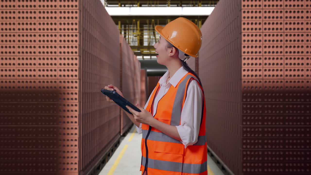Side View Of Asian Female Engineer With Safety Helmet Taking Note On The Tablet And Looking Around While Standing With Red Brick Packed in Stacks Are Stored