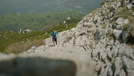 Hiker-with-hiking-poles-in-blue-shirt-climbing-up-on-sharp-rocks-towards-the-camera,-in-background-is-green-karst-valley