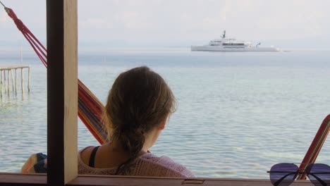 Woman-relaxes-on-Kri-Island-in-the-Raja-Ampat-archipelago,-Indonesia,-gazing-at-a-distant-luxurious-yacht