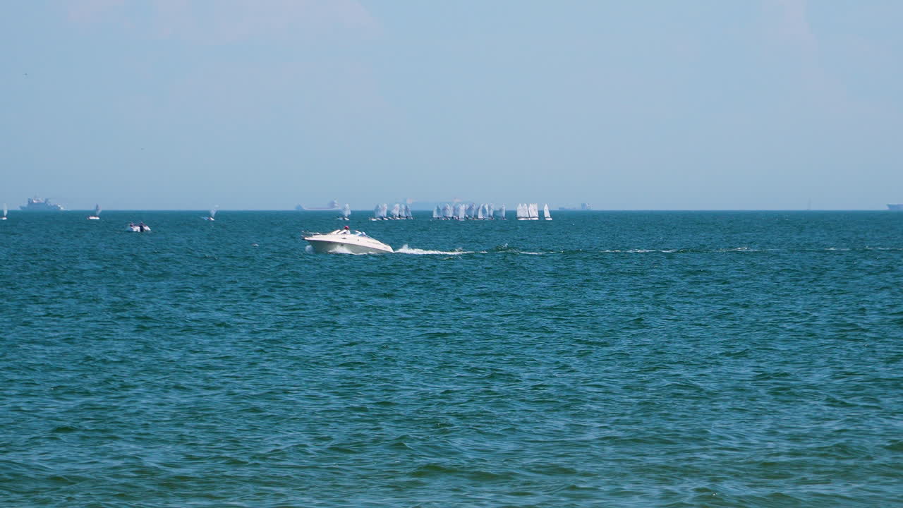 A speeding powerboat cutting through the blue sea, leaving a wake, with dozens of sailboats and windsurfers visible in the distance on a sunny day