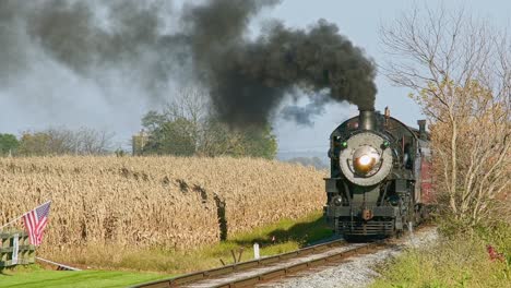 a view of an approaching antique steam passenger train, blowing black smoke, passing corn fields