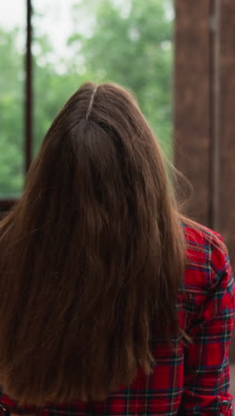 woman walks tilting head back along terrace. lady with long red hair wearing checkered shirt rests in studio with panoramic windows. luxury house