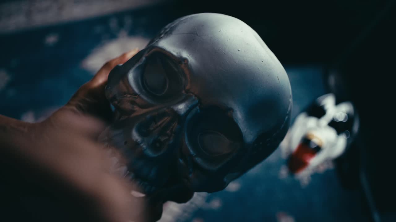 Close-up footage of human hands holding and examining a metallic skull model in dramatic low light. The shot emphasizes texture, shadows, and detail, creating a moody and cinematic atmosphere