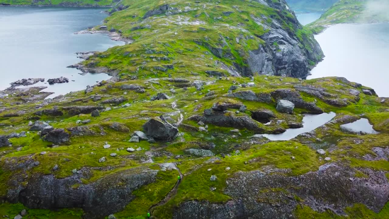 Aerial footage flying over moss covered rocky terrain and mountain landscape in Lofoten Norway while two hikers are walking below. Two large blue colored lakes on both side of the mountains. Mist fog.