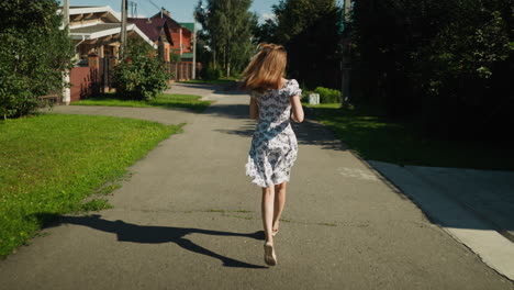 back view of young woman running along quiet residential street as her hair sways with motion, bright daylight highlights her floral dress while surrounding homes and greenery frame her path home