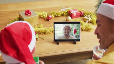 padre y hijo caucásicos con sombreros de santa usando una tableta para una videollamada de navidad con un hombre en la pantalla