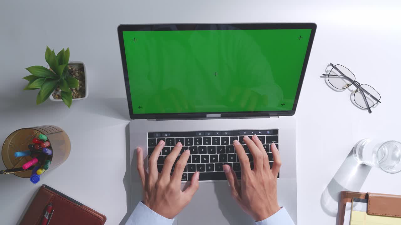 vista de arriba hacia abajo de una computadora portátil con pantalla verde simulada pantalla chromakey en un escritorio de oficina de madera al lado de la libreta con bolígrafos, gafas y un vaso de agua.