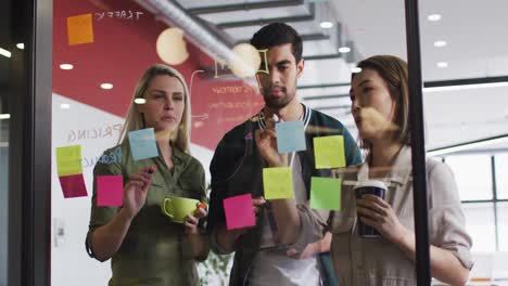 Diverse-Bürokollegen-Schreiben-Auf-Einer-Glastafel-Und-Diskutieren-Gemeinsam-Im-Modernen-Büro