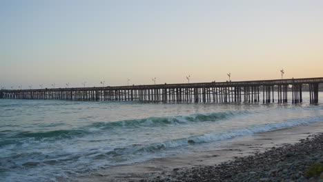 Toma-Panorámica-Del-Muelle-De-Ventura-Con-Olas-Del-Océano-Pacífico-Rodando-Hacia-La-Orilla-Con-Una-Brillante-Puesta-De-Sol-Naranja-Reflejada-En-El-Agua-Ubicada-En-El-Sur-De-California