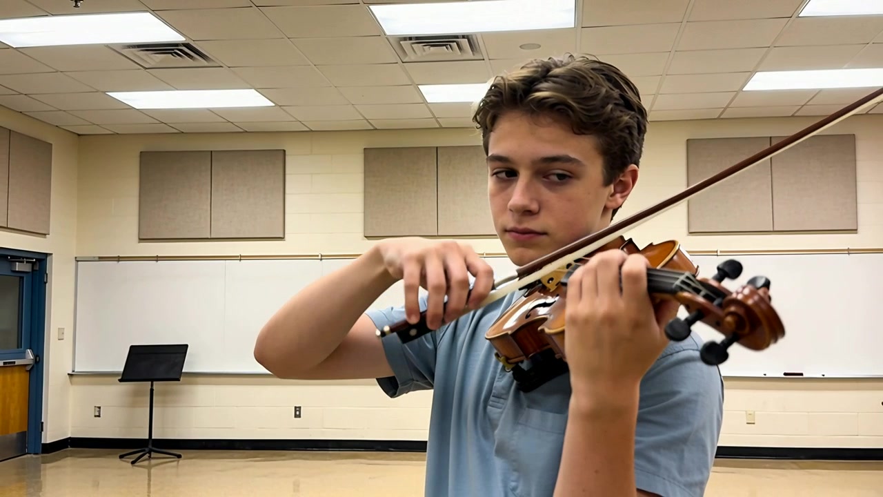 Starting practice teen boy in light blue polo bowing violin and fingering notes in music room