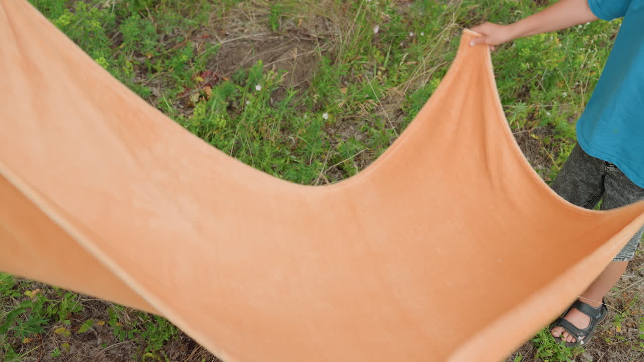 Boy arranges colorful cloth outside, Juvenile child neatly organizing fabric in bright outdoor landscape, Youthful boy carefully positioning colorful blanket amid vibrant sunlit grass field