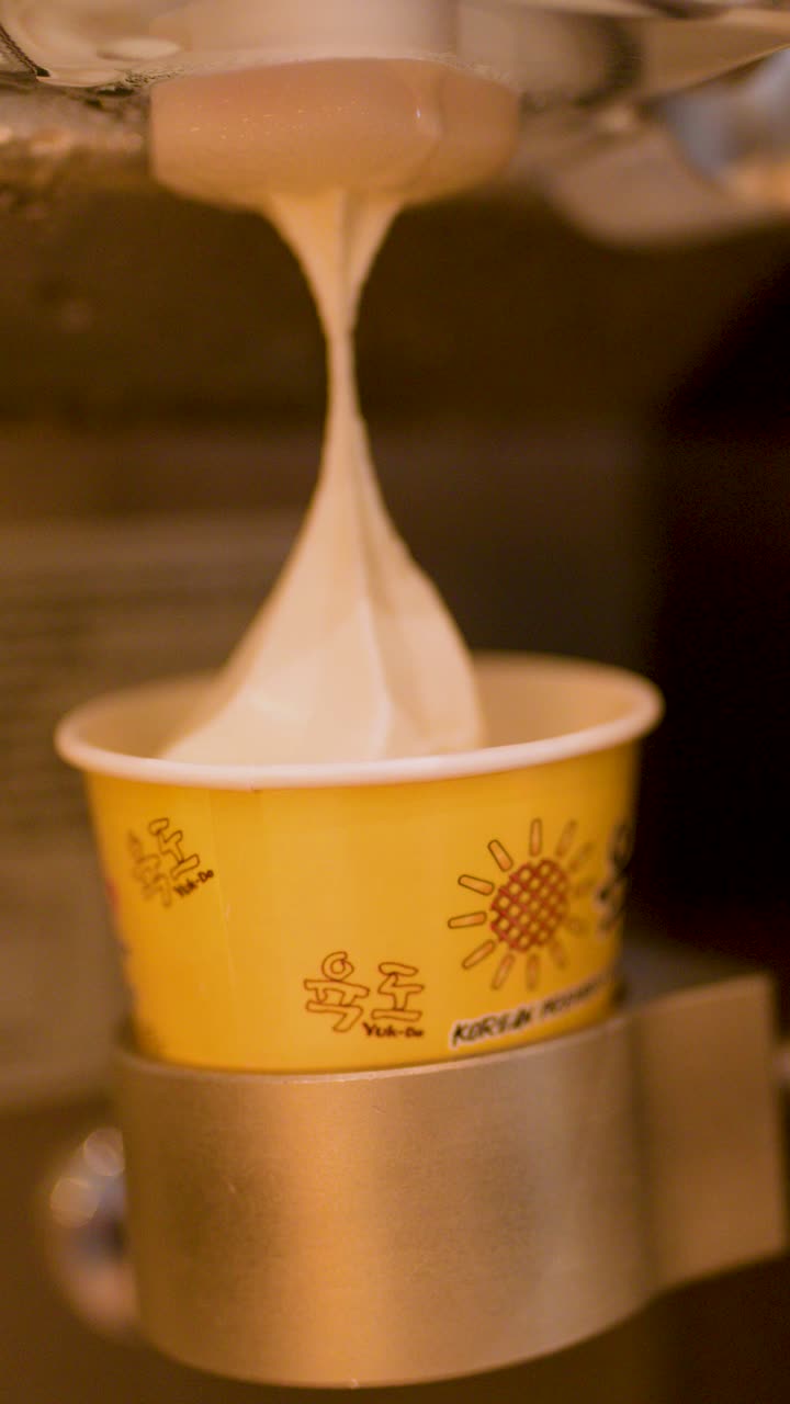 Soft serve ice cream dispensed into yellow paper cup by machine, warm lighting, close-up view