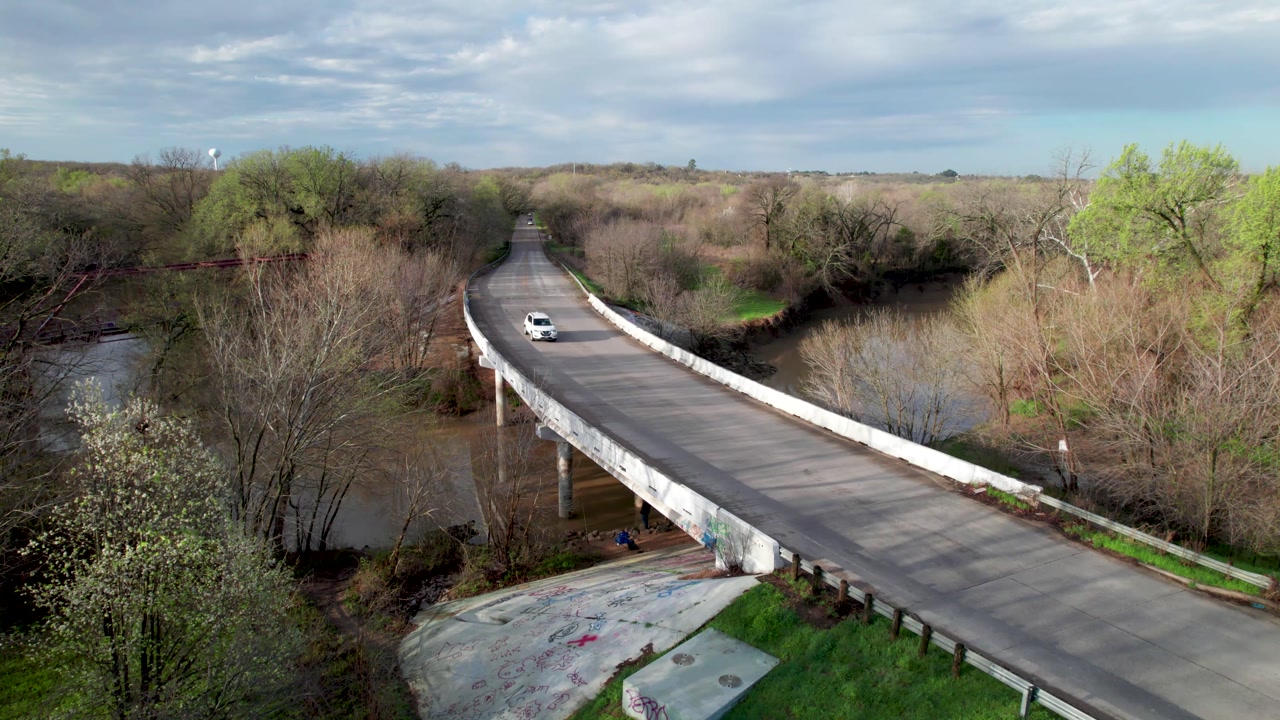 Static aerial video of cars driving on Old Alton Road near the Goatman Bridge in Lantana, Texas. Several cars can be seen driving on the road.