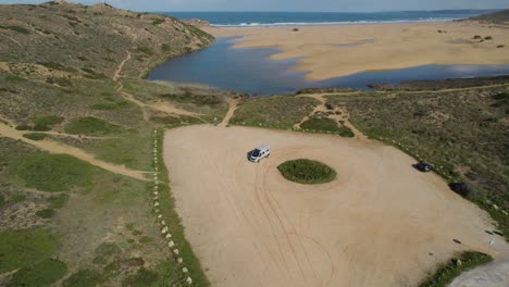 Drone-flight-flowing-wan-car-starting-from-sandy-ground-surface-and-getting-on-the-road,-recorded-at-Bordeira-Portugal-hillside-coastline-overlooking-seascape-