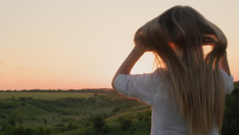 a teenage girl touches her long blond hair, stands against the backdrop of a picturesque landscape at sunset. back view