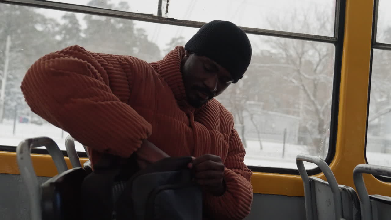 Uomo di colore che apre lo zaino sull'autobus, con maglione arancione e berretto di lana, cercando biglietto e portafoglio, mani concentrate ed espressione intenta, città innevata visibile attraverso il finestrino, un momento spontaneo del mattino.