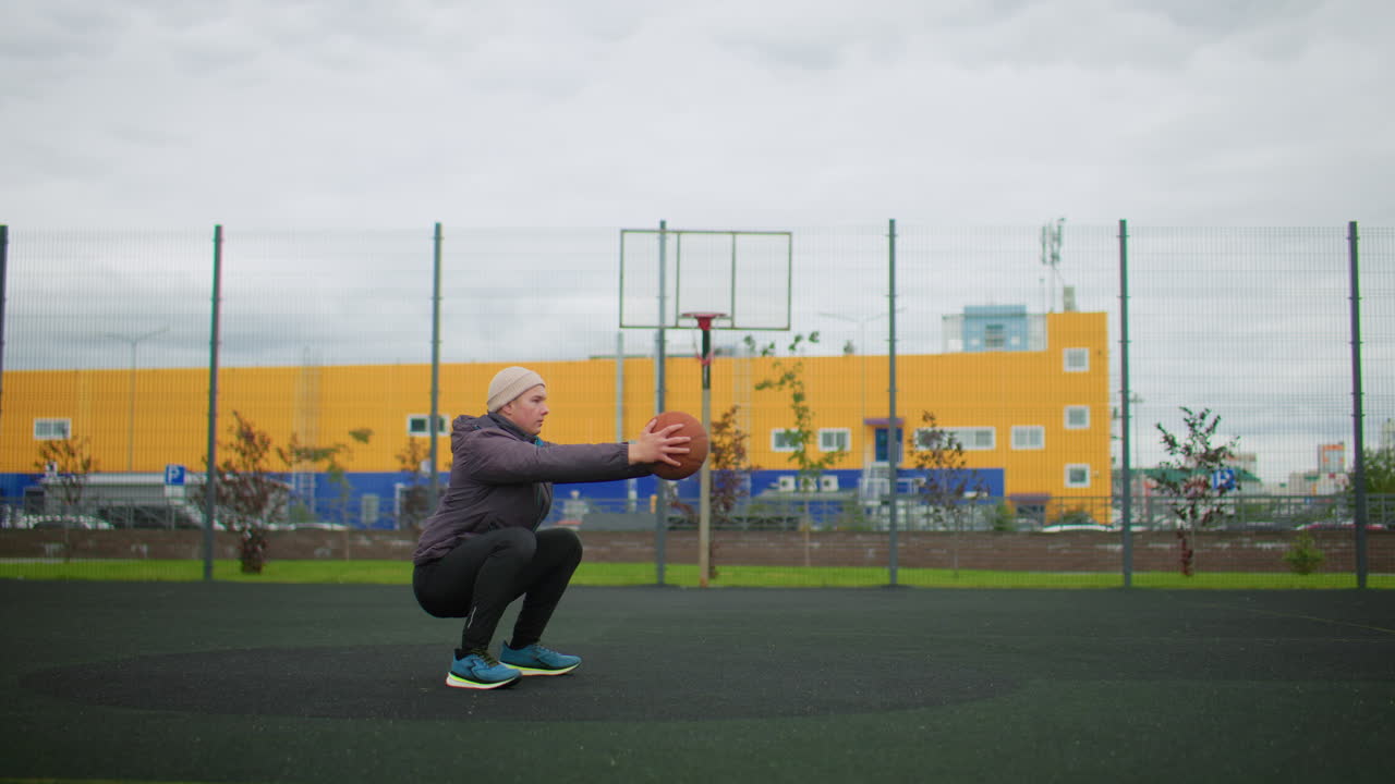Allenamento di basket all'aperto su un campo recintato, un atleta solitario con felpa e berretto esegue squat e passaggi al petto sotto un cielo nuvoloso accanto a un edificio industriale giallo, allenamento mirato con determinazione