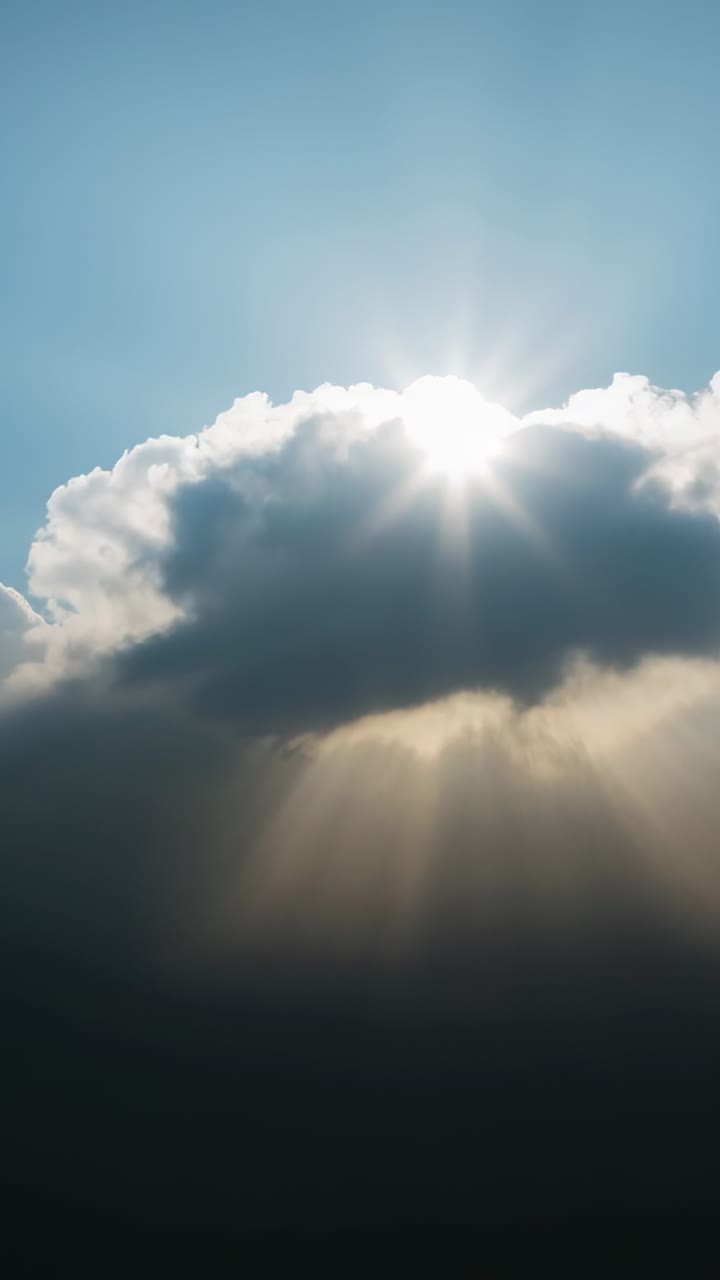 Vertical video: Sun peeking behind large cumulus cloud, casting starburst and sunbeams in blue sky