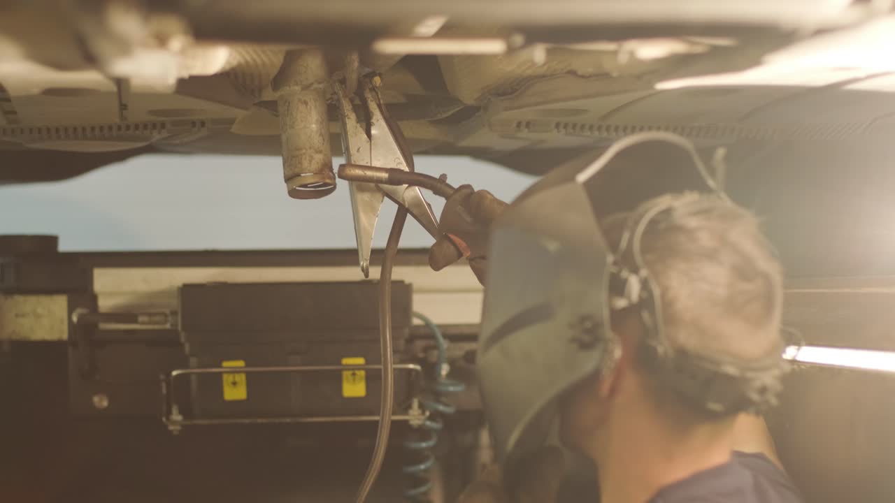 trabajador de servicio de automóviles soldas y repara una tubería de escape de un automóvil. trabajo de soldadura de primer plano en un automóvil levantado. vista desde la parte trasera. mecánico soldas la parte inferior del automóvil.