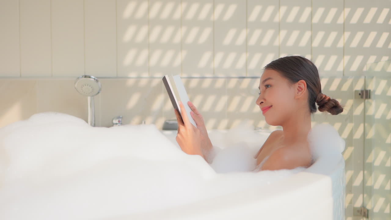Woman relaxing and reading in bathtub filled with bubbles. Young Asian lady taking a bubble bath and enjoying some quiet time with a book. Patterns of sunlight on wall near girl bathing