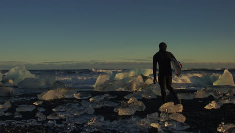 a surfer walks along the water’s edge, stepping carefully between scattered ice formations
