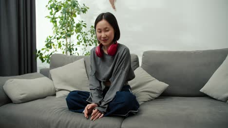 Portrait-of-gentle-asian-girl-wearing-wireless-headphones,-sitting-on-couch-and-smiling-to-the-camera