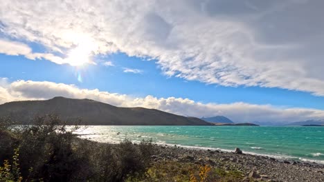 Wide-angle daytime pan reveals Lake Tekapo’s turquoise waters, rocky shoreline, and distant snow-capped mountains under striking, dynamic cloud formations with natural sunlight