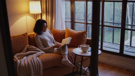 woman reading a book on a cozy sofa during a rainy day