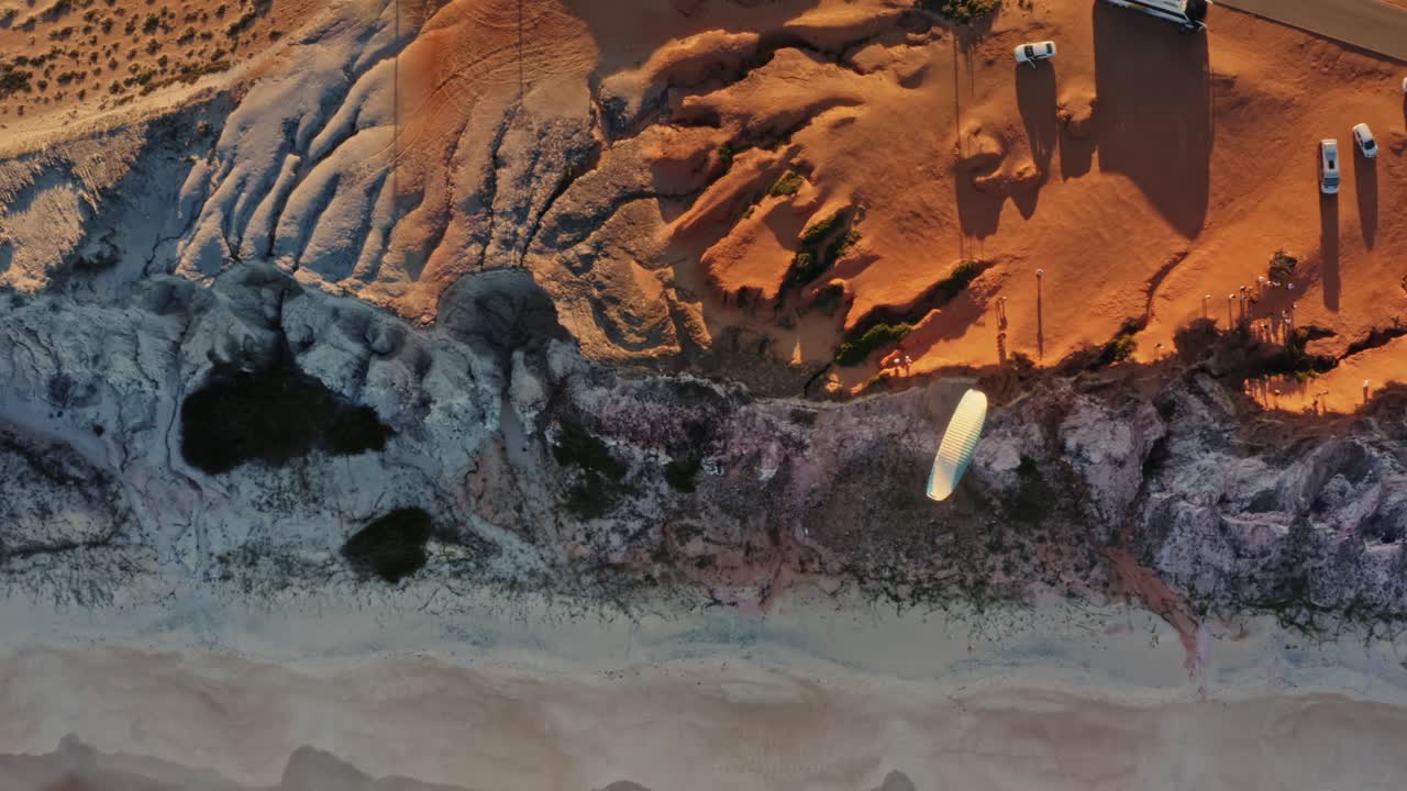 Aerial drone bird's eye top view following a paraglider flying over the Cliffs of Cacimbinhas in Pipa, Brazil Rio Grande do Norte with golden sand below and colorful orange cliffs above