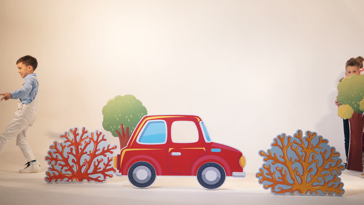 Cute little boy in white trousers comes to the children's room and shows surprised gestures. Childish studio with decor of toyful car, trees and bushes and kids playing in it.