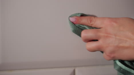 close up hand view of lady wearing beads cleaning smooth white kitchen wall with green cloth focusing on hygiene neatness home care maintenance