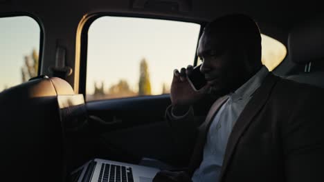 hombre de negocios confiado con color de piel negra con barba en una chaqueta marrón se comunica en un teléfono inteligente y trabaja en una computadora portátil mientras conduce en el asiento del pasajero en un coche moderno fuera de la ciudad