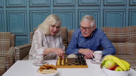 Retired-senior-couple-talking-drinking-tea,-playing-chess-in-modern-living-home-room-lounge-together