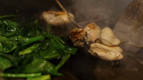 Close-up view of oysters and fresh greens being stir-fried on a steaming hot wok.