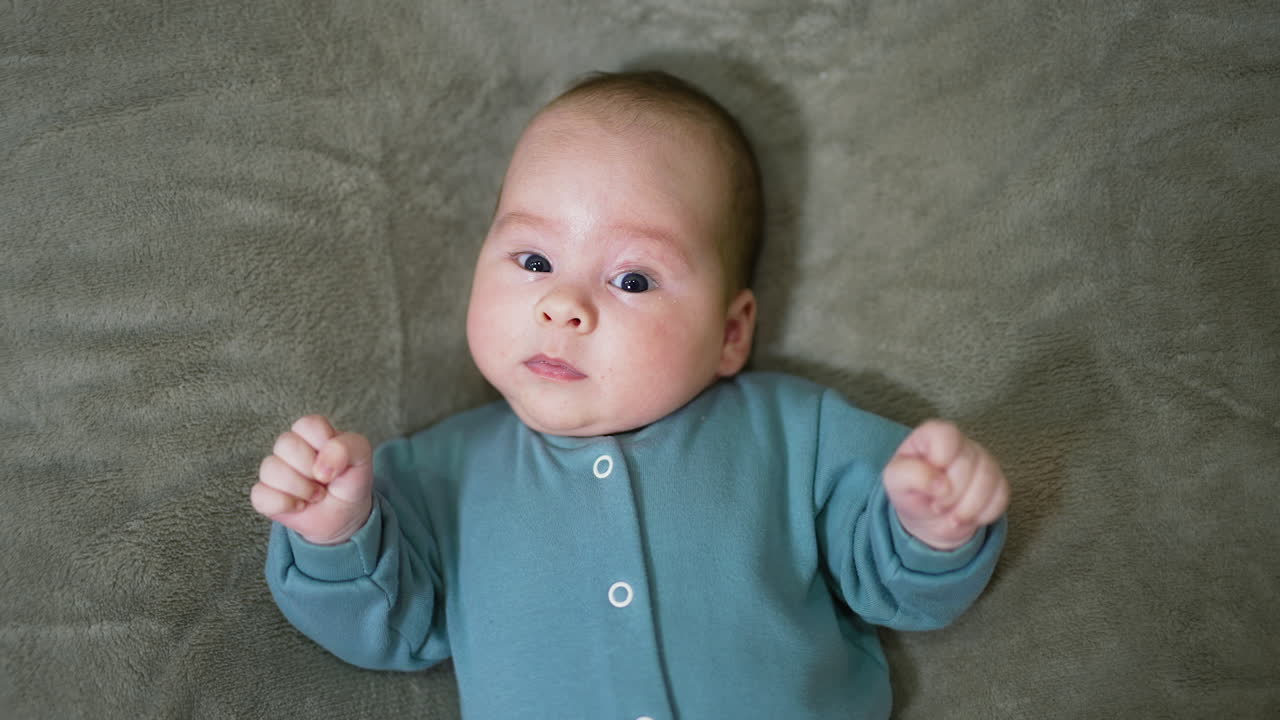 Baby boy in blue sweater lies and looks straight ahead. Little child watches camera with surprise and lies still. Top view. Close up.