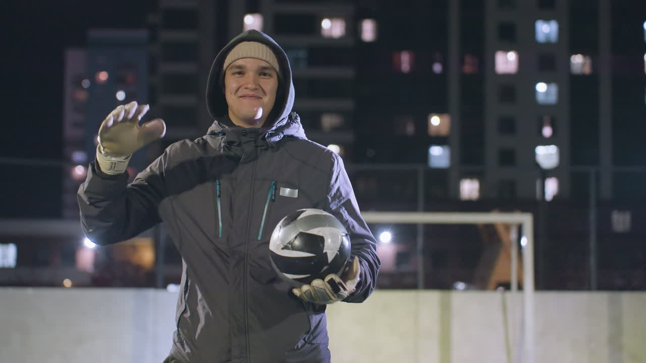 hombre sonriente con capucha y guantes golpeando casualmente una pelota de fútbol en su mano bajo luces nocturnas, entorno deportivo urbano con edificios de gran altura y fondo bokeh