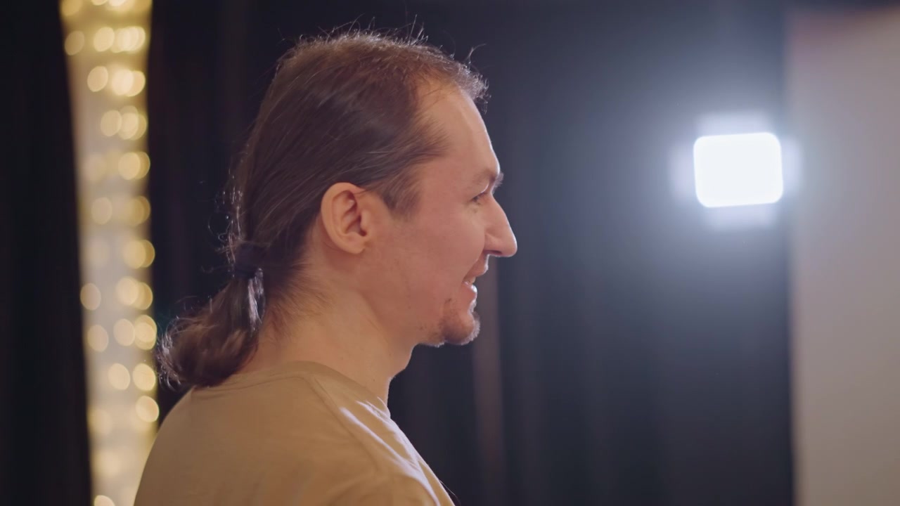 Coach di musica in prova in studio, di profilo e concentrato sotto luci calde, con uno sfondo di tenda scura. Con una coda di cavallo, fa un gesto espressivo verso uno studente non visibile, in un'atmosfera istruttiva che guida la tecnica vocale.
