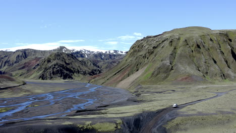 aerial view of a 4x4 white car driving on a dirt road by a wide riverbed and range of green and grey mountains in the background in iceland near thakgil campsite