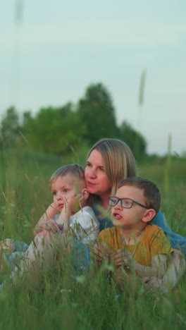 a mother in a blue gown sits in a lush, green field with her two children, the older one in a yellow top and glasses, and the younger one in a white top. they are gazing at their surroundings