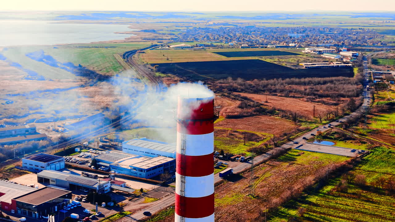 Vista aerea ravvicinata di una ciminiera a strisce rosse e bianche che emette fumo, con una città e campi sullo sfondo. Ciminiera industriale con fumo su un paesaggio rurale.