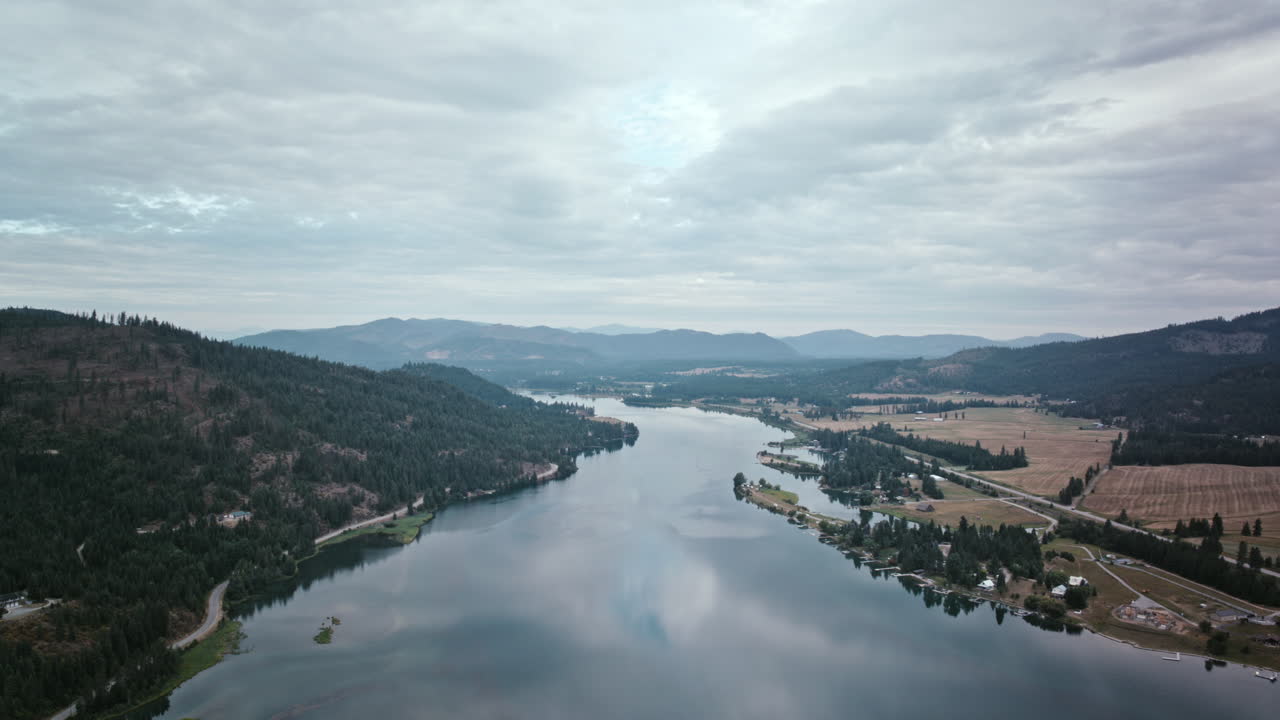 Aerial video of the Priest River under an overcast sky, framed by green mountains and open fields, the water stretching toward the horizon