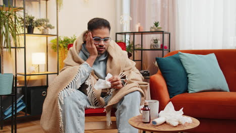 sick ill indian man suffering from cold or allergy sits on home chair sneezes wipes snot into napkin
