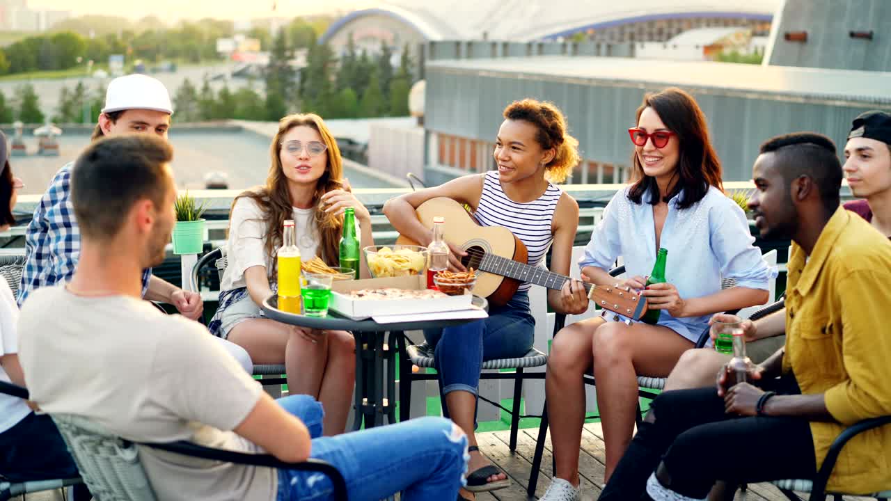 African American girl is playing the guitar while her friends multiethnic group are singing and moving hands enjoying party on rooftop. Table with food and drinks is visible.