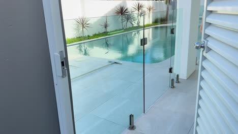 a man in a blue shirt unlocks and opens a white louvered gate, entering a modern outdoor patio with a glass fence and swimming pool in daylight