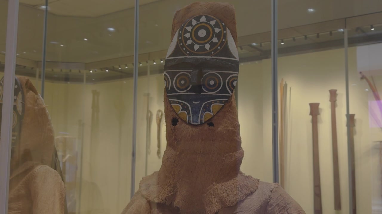 Geometrik bir maske içeren Amazon ritüel giysisi, cam bir müze vitrininde sergileniyor. Kültürel belgeseller veya eğitim içerikleri için idealdir.